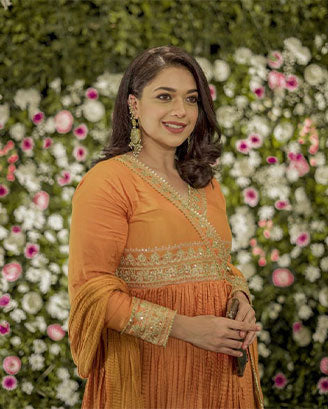 Woman wearing an orange traditional outfit with embroidered details, standing in a floral garden setting