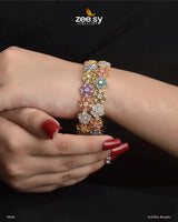 Achillea Bangles, a traditional kangan set featuring intricate floral patterns, adorned with uncut white stones and vibrant accent gems. Embellished with dazzling American cut diamond stones and finished with a rhodium polish, these handcrafted kadas exude elegance, making them ideal for weddings, bridal wear, and festive occasions.