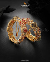 Opulenza Bangles radiate grandeur with their intricate floral motifs and regal design. Embellished with vibrant stones and delicate filigree work, these bangles exude luxury and tradition. Featuring an elegant hand chain attachment, they make the perfect statement piece for bridal and festive occasions.