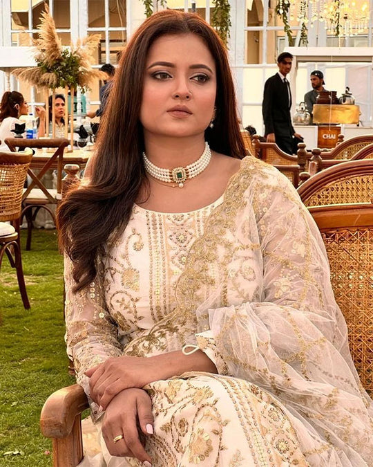 Woman wearing an elegant cream embroidered traditional outfit with matching jewelry, seated in an outdoor garden setting