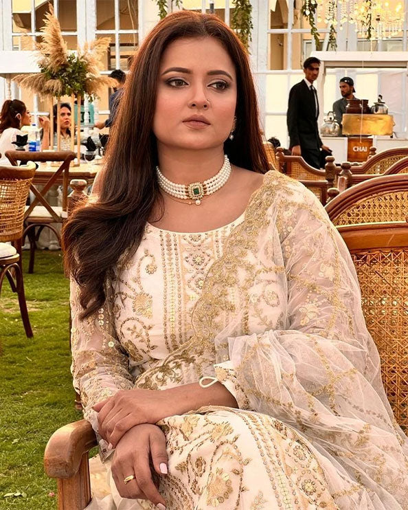 Woman wearing an elegant cream embroidered traditional outfit with matching jewelry, seated in an outdoor garden setting